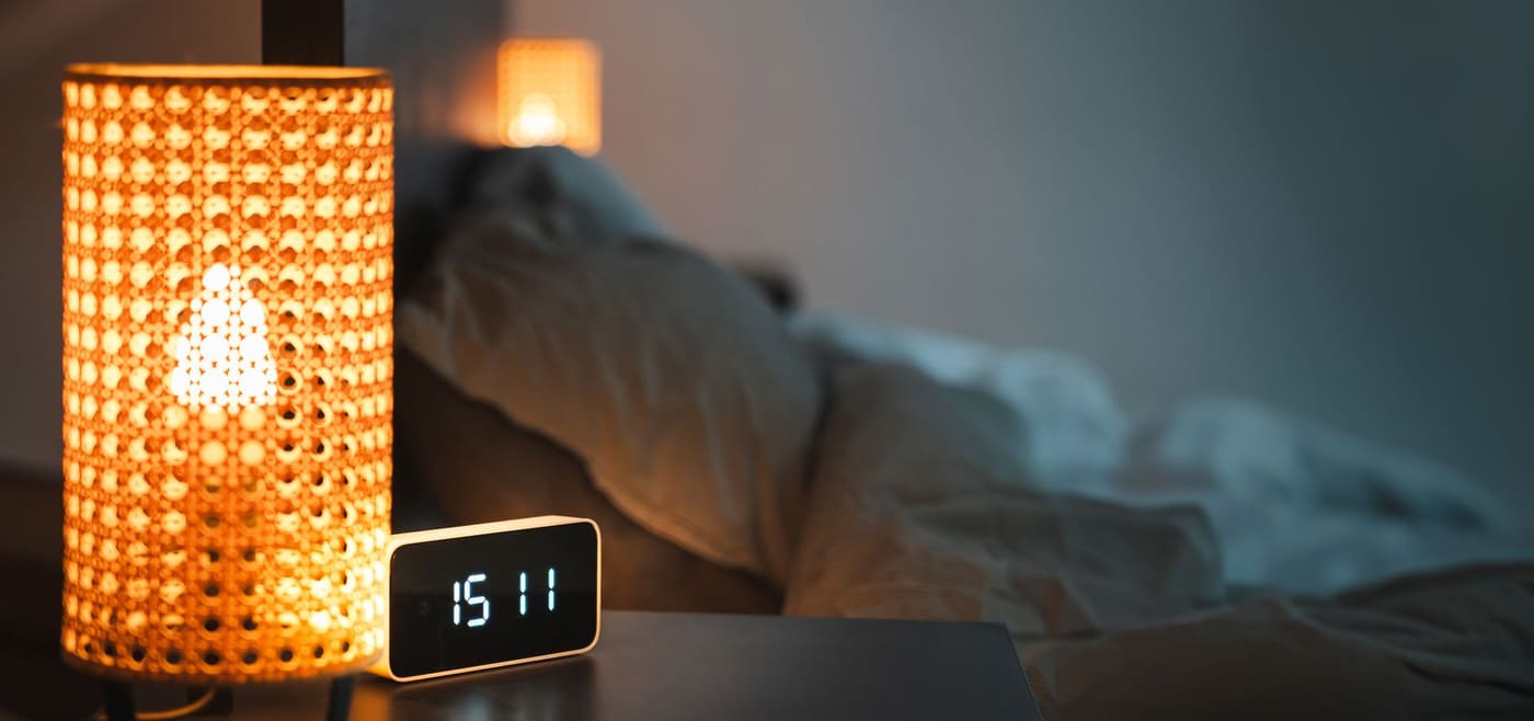 A bedside lamp glowing next to a digital alarm clock in a dim bedroom