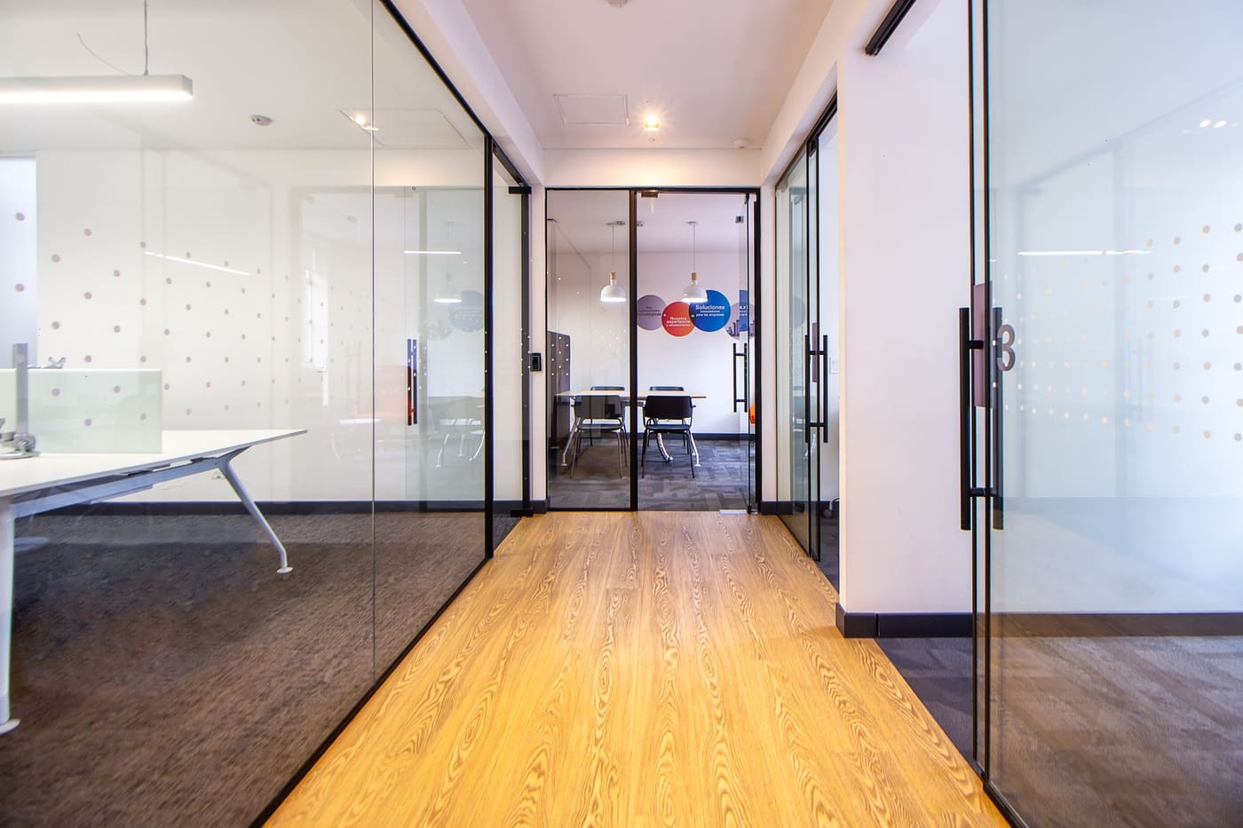 A bright office hallway with glass walls and a wood floor leading toward a meeting room