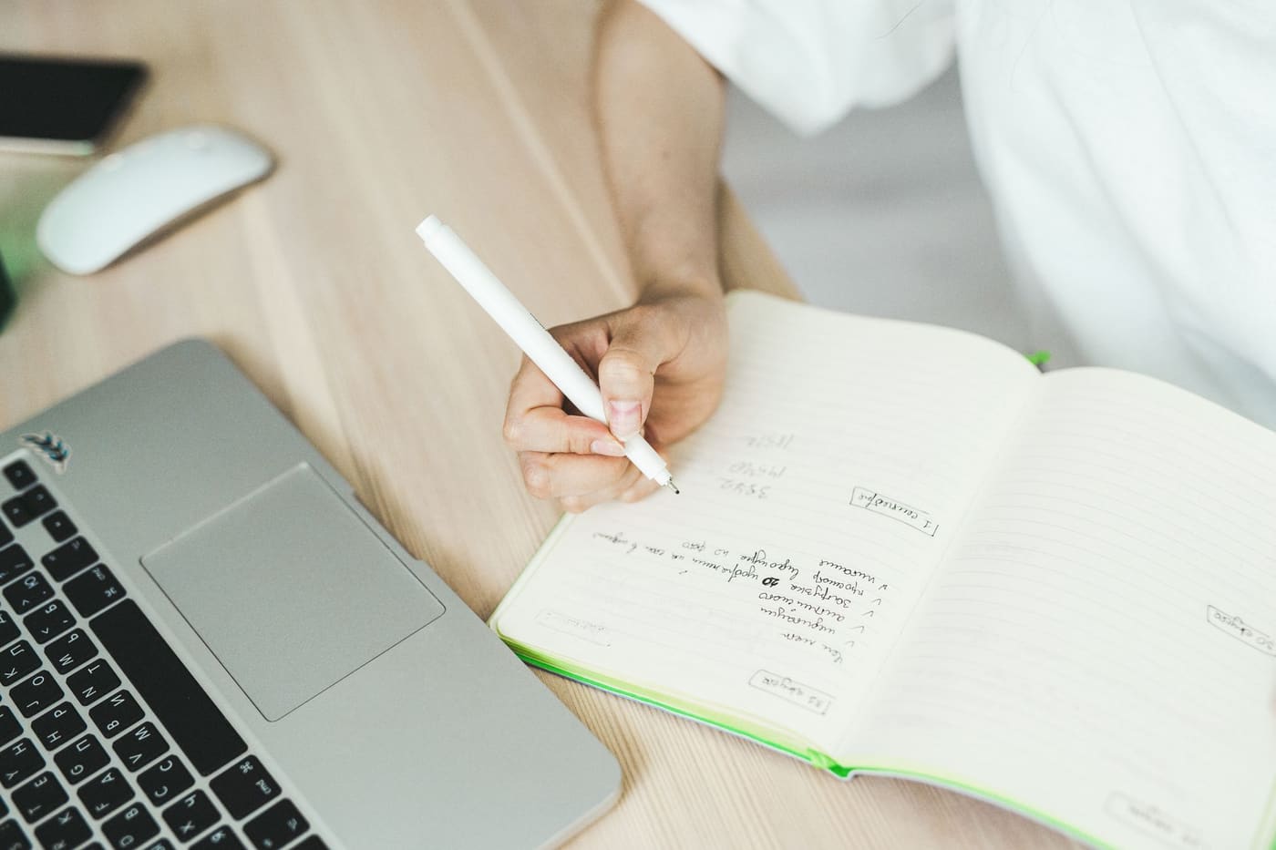 A hand writing in an open notebook next to a laptop on a light wooden desk