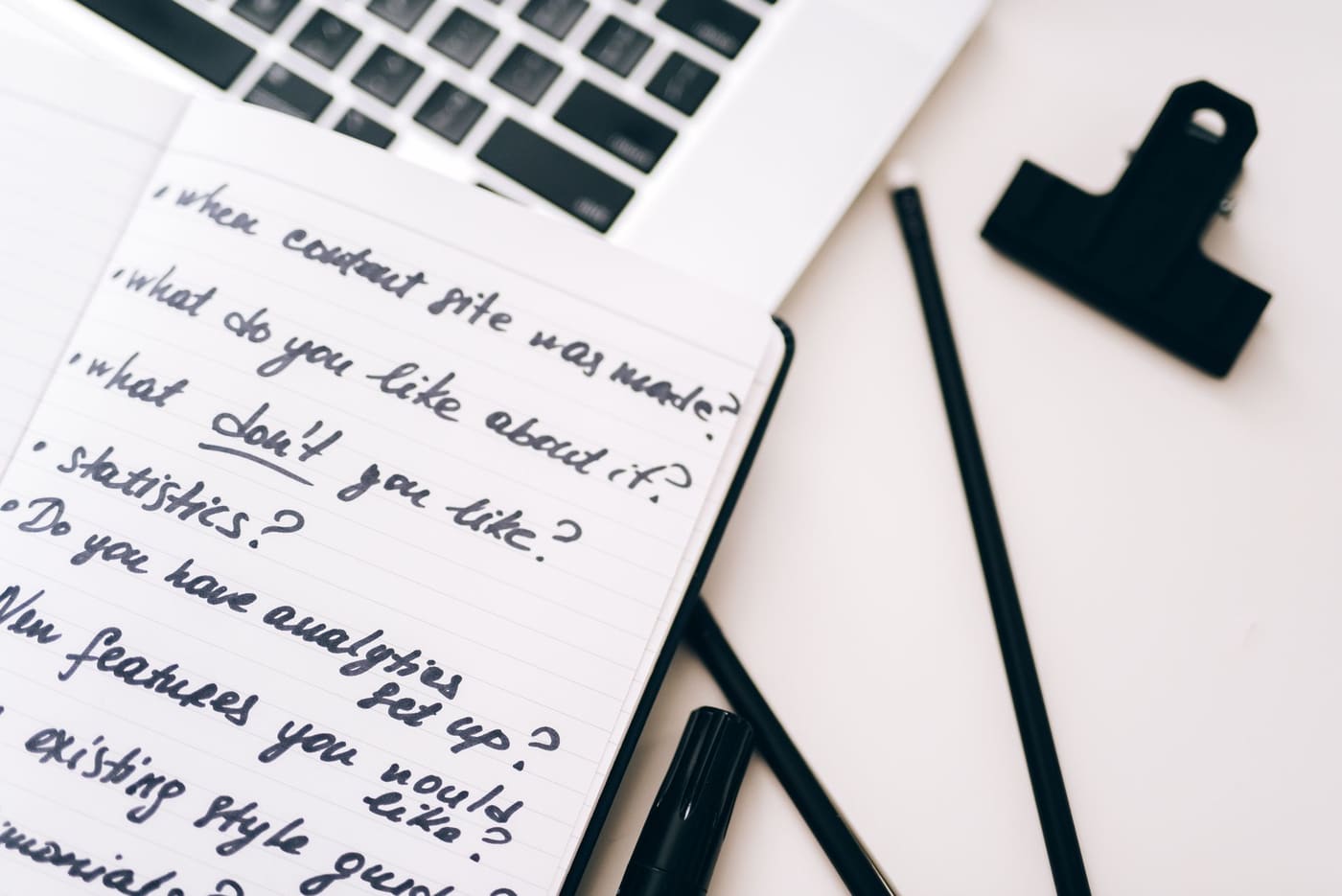 A notebook page of handwritten questions beside a laptop and a black pen