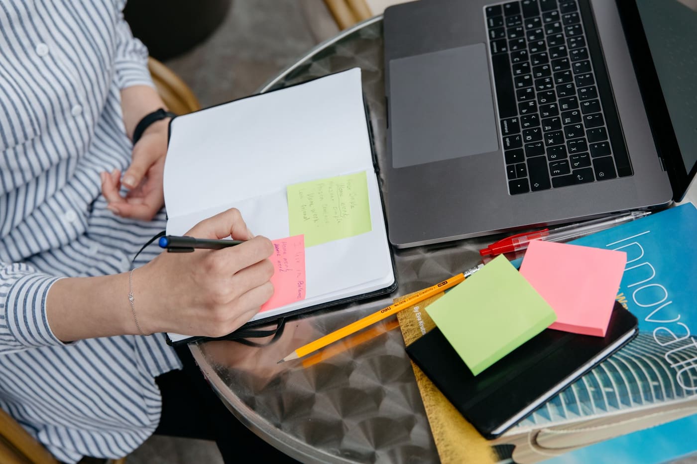 A person writing in a notebook beside a laptop and colorful sticky notes on a desk