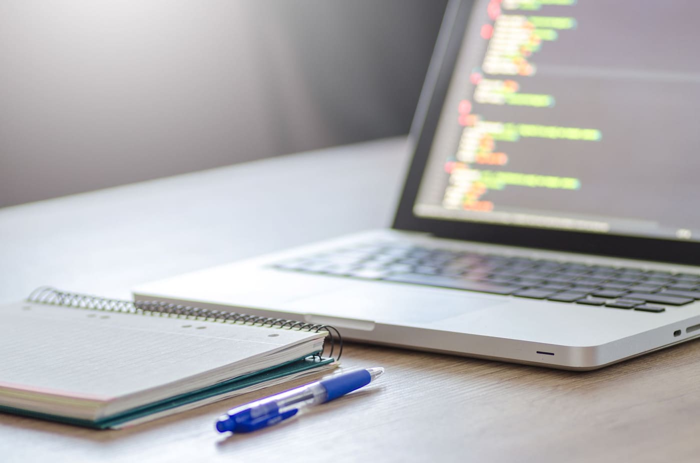 A laptop with code on screen next to a spiral notebook and a blue pen