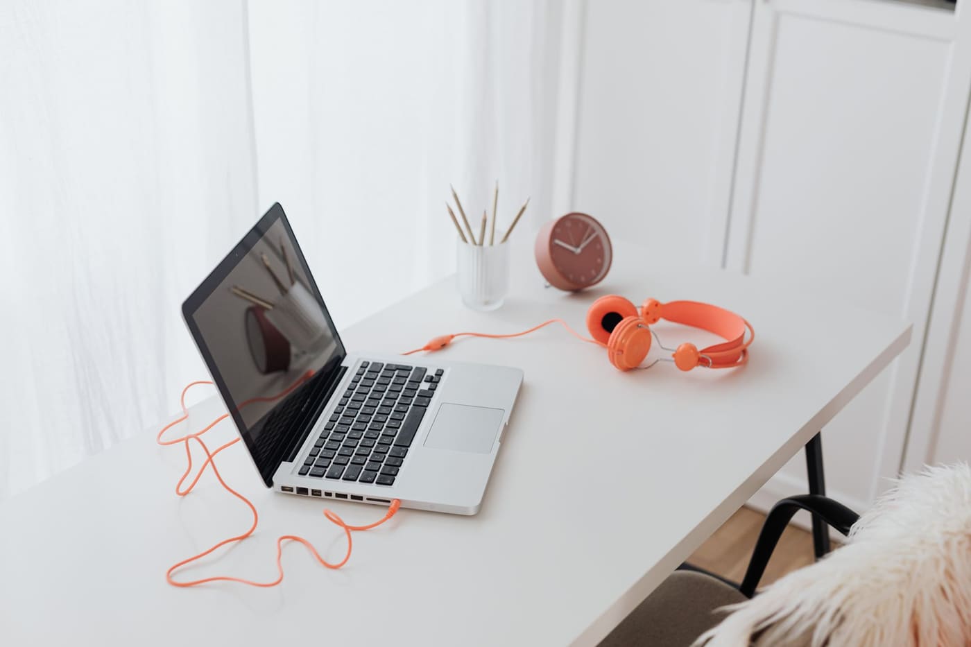 A laptop and orange headphones on a clean white desk by a window