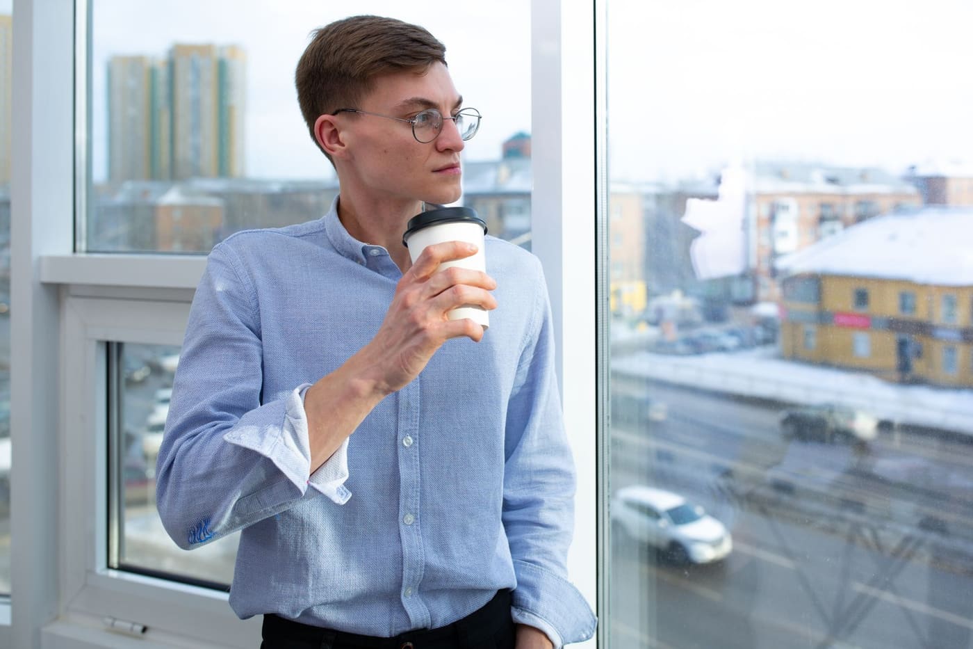 A man with glasses holding a cup beside a large window overlooking a city street