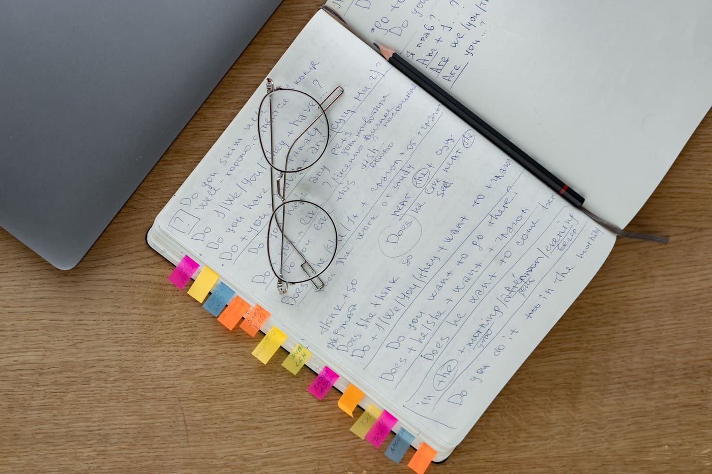 An open notebook with handwritten notes, glasses and colored index tabs on a wooden desk