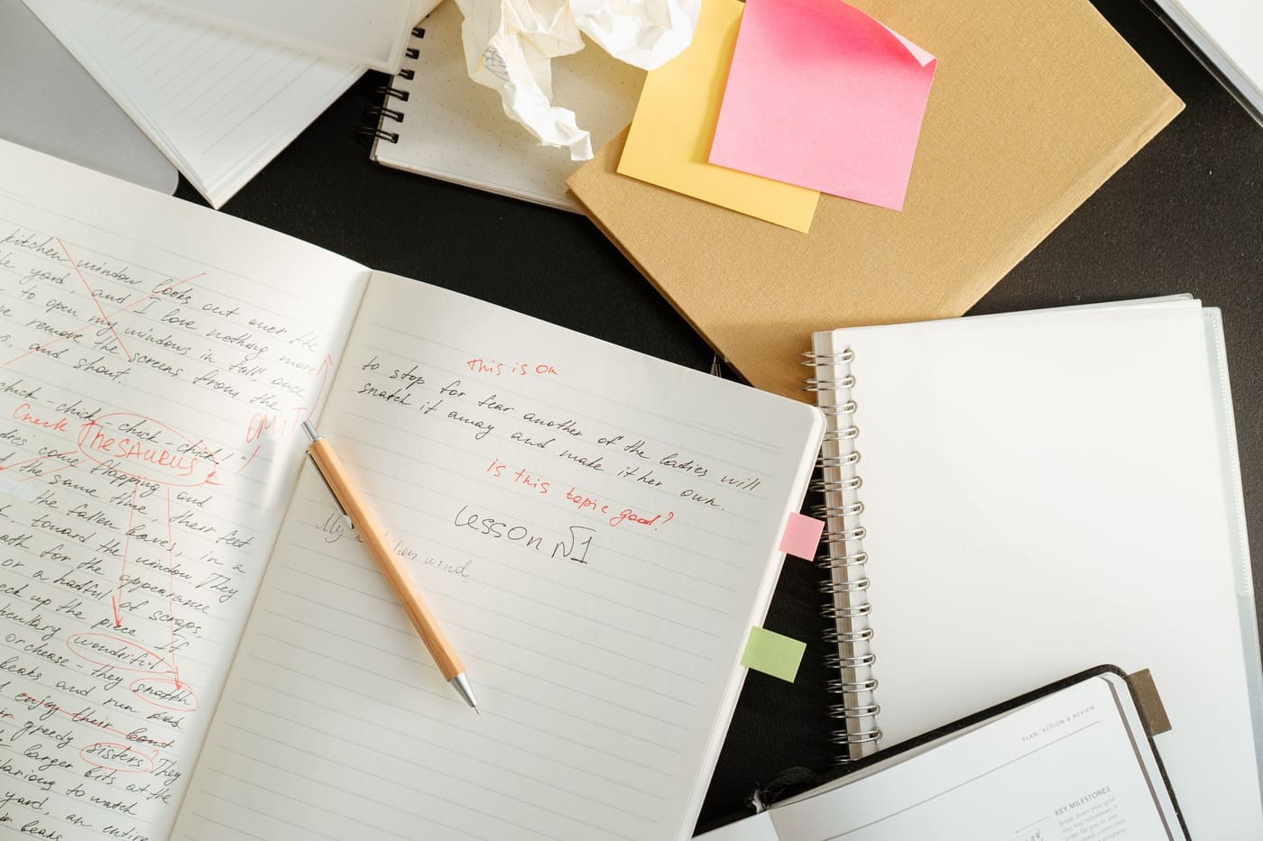 An open notebook with handwritten notes and red corrections, with sticky notes and a pencil beside it