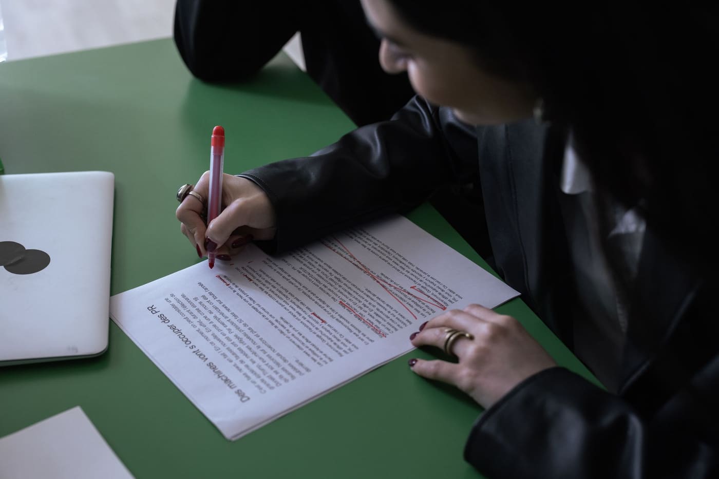 A person marking up a printed page with a red pen at a desk with a laptop
