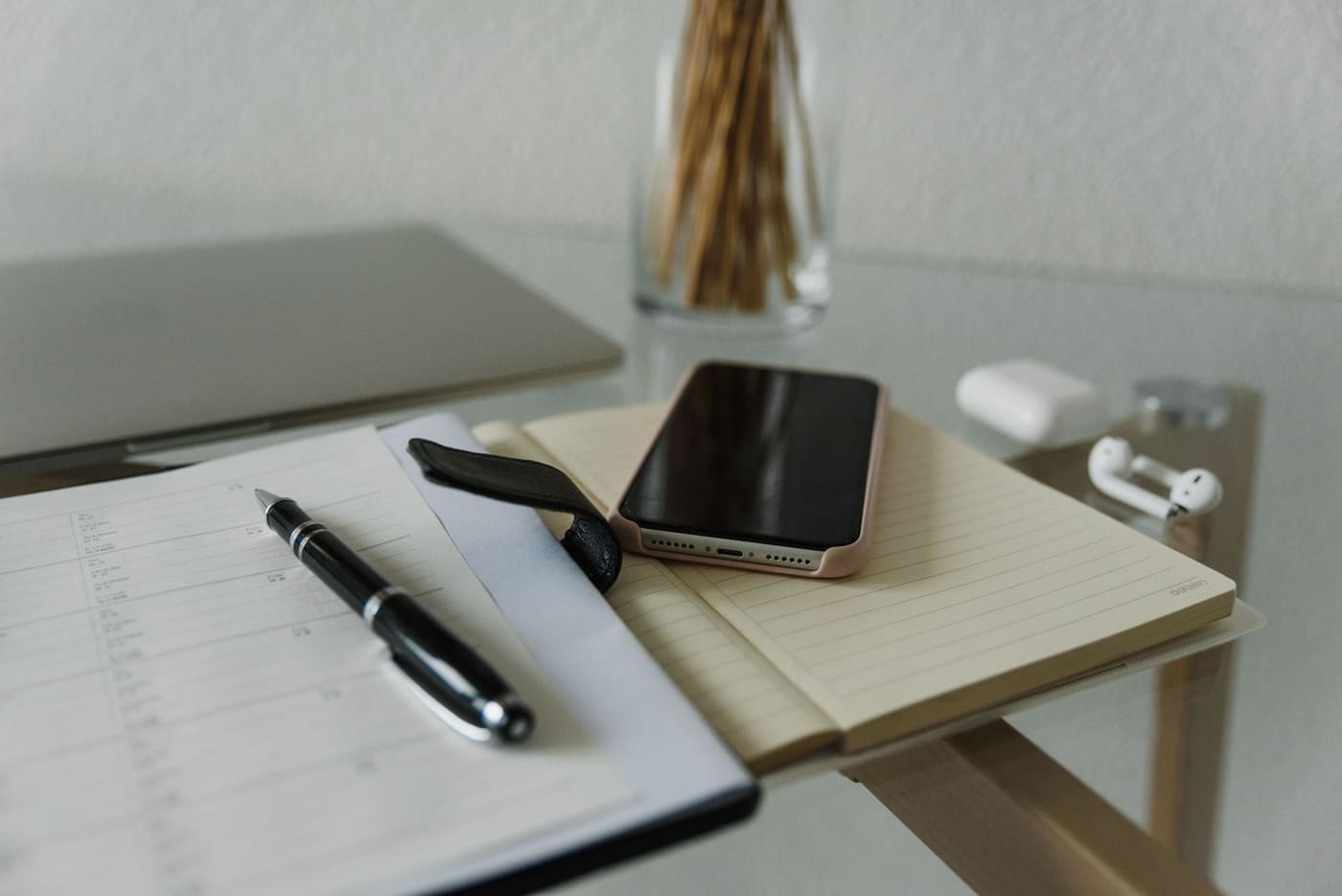 A smartphone, a notebook with a pen and wireless earbuds on a glass desk