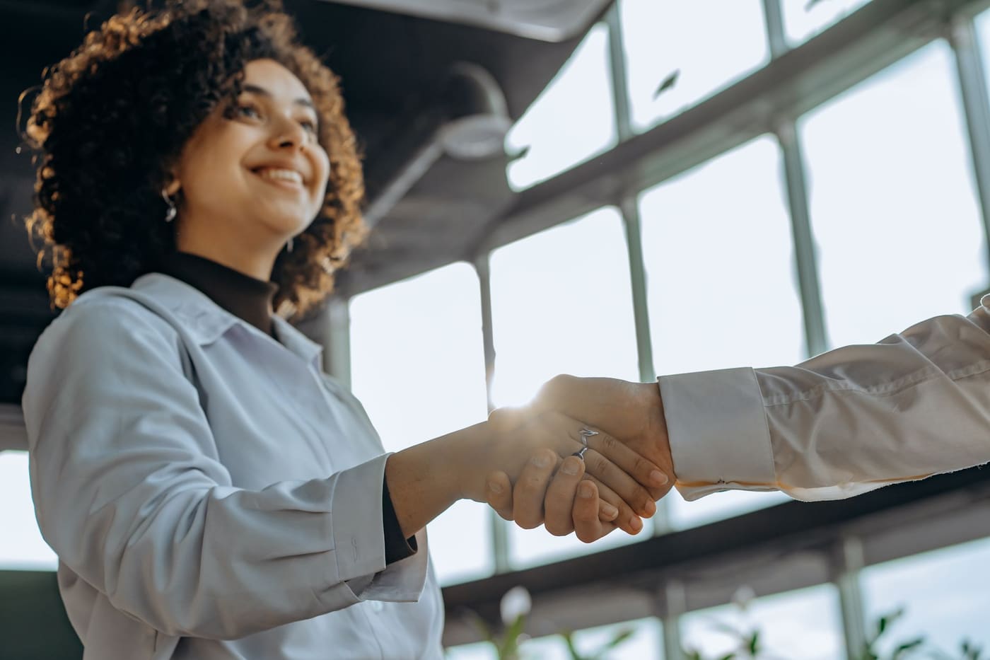 A smiling woman shaking hands with someone in a bright office with large windows