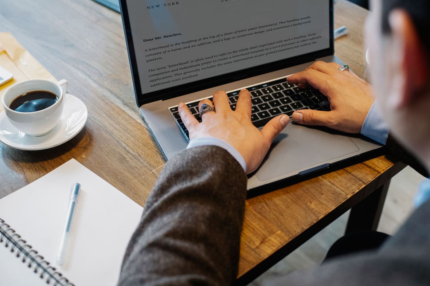 A person typing on a laptop at a wooden desk with a cup of coffee and a notebook