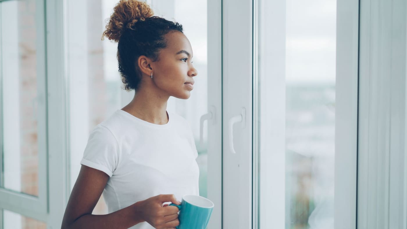 A woman holding a mug and looking calmly out of a bright window