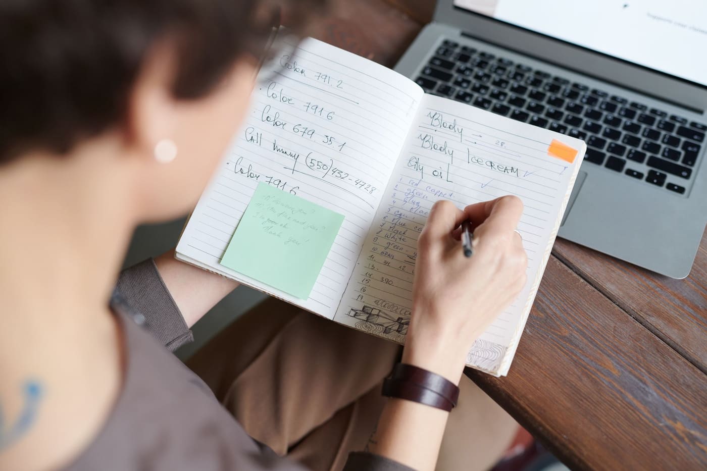 A person writing in a notebook next to an open laptop on a wooden table